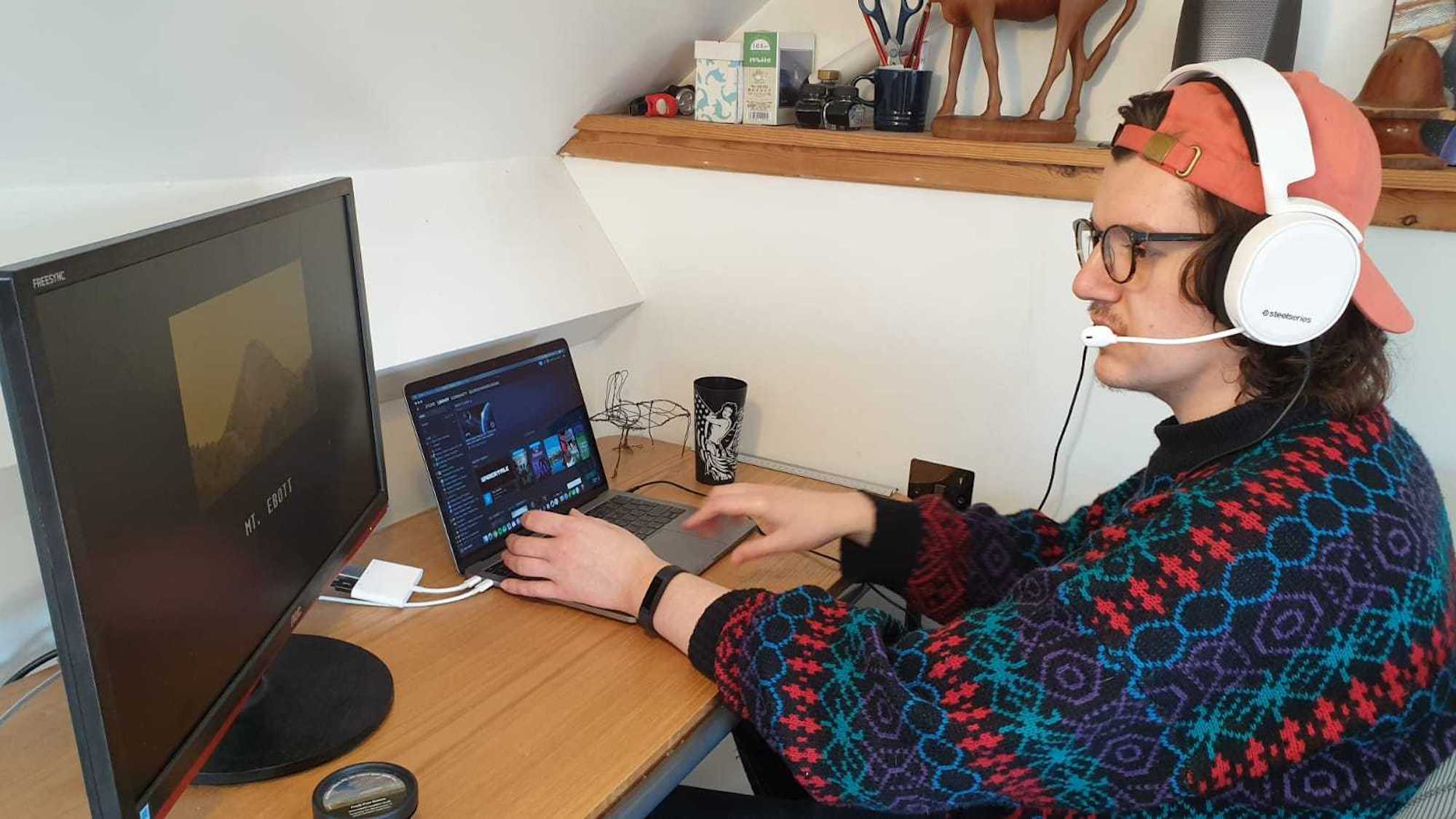 Photo of a man wearing a jumper and baseball cap playing video games on a laptop and monitor