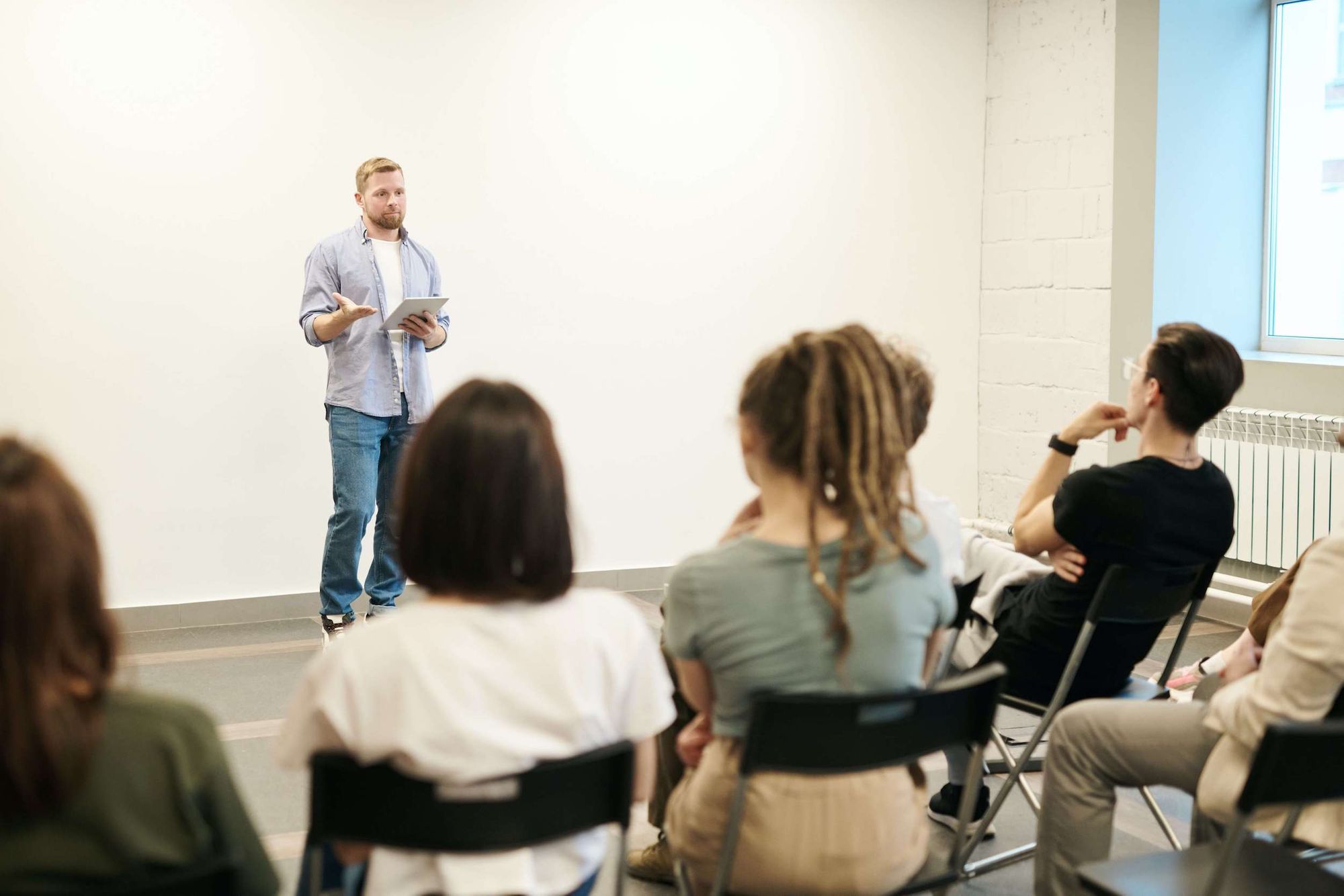 man holding iPad speaking to a group of people seated on plastic chairs