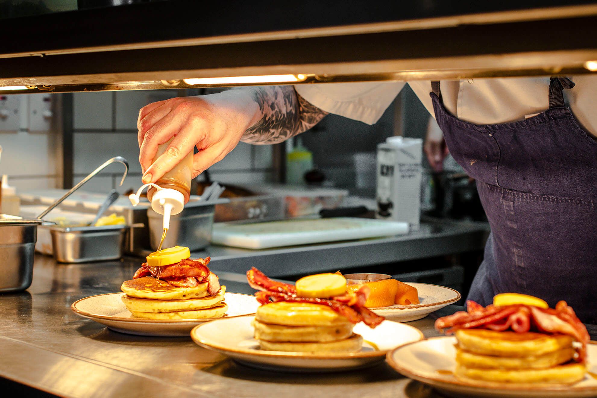 Pancakes and bacon being prepared in by chef in cafe kitchen.