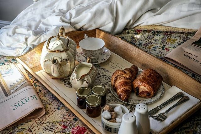 Teapot, croissants and jam on a tray, beside a newspaper, on an embroidered bedspread