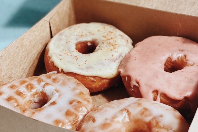 A cardboard box of four glazed donuts: two traditional glaze, one yellow, and one pink