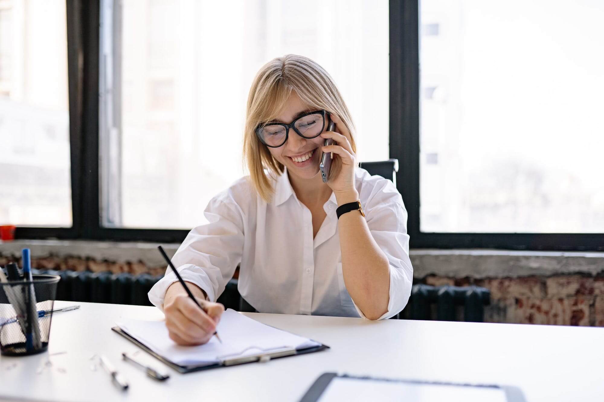 Smartly dressed woman with blonde hair talking on a mobile phone while taking notes.