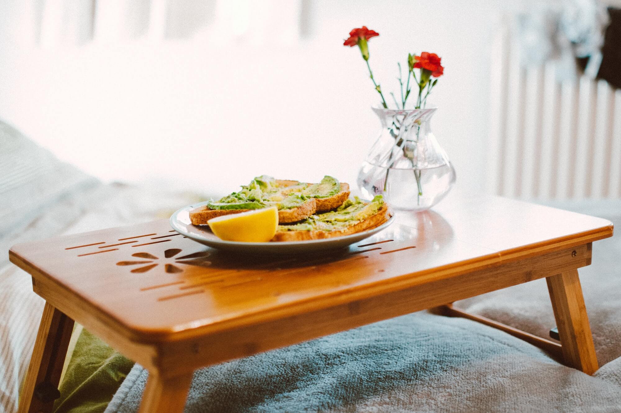 Avocado on toast served on a tray with flowers in a vase atop a bed