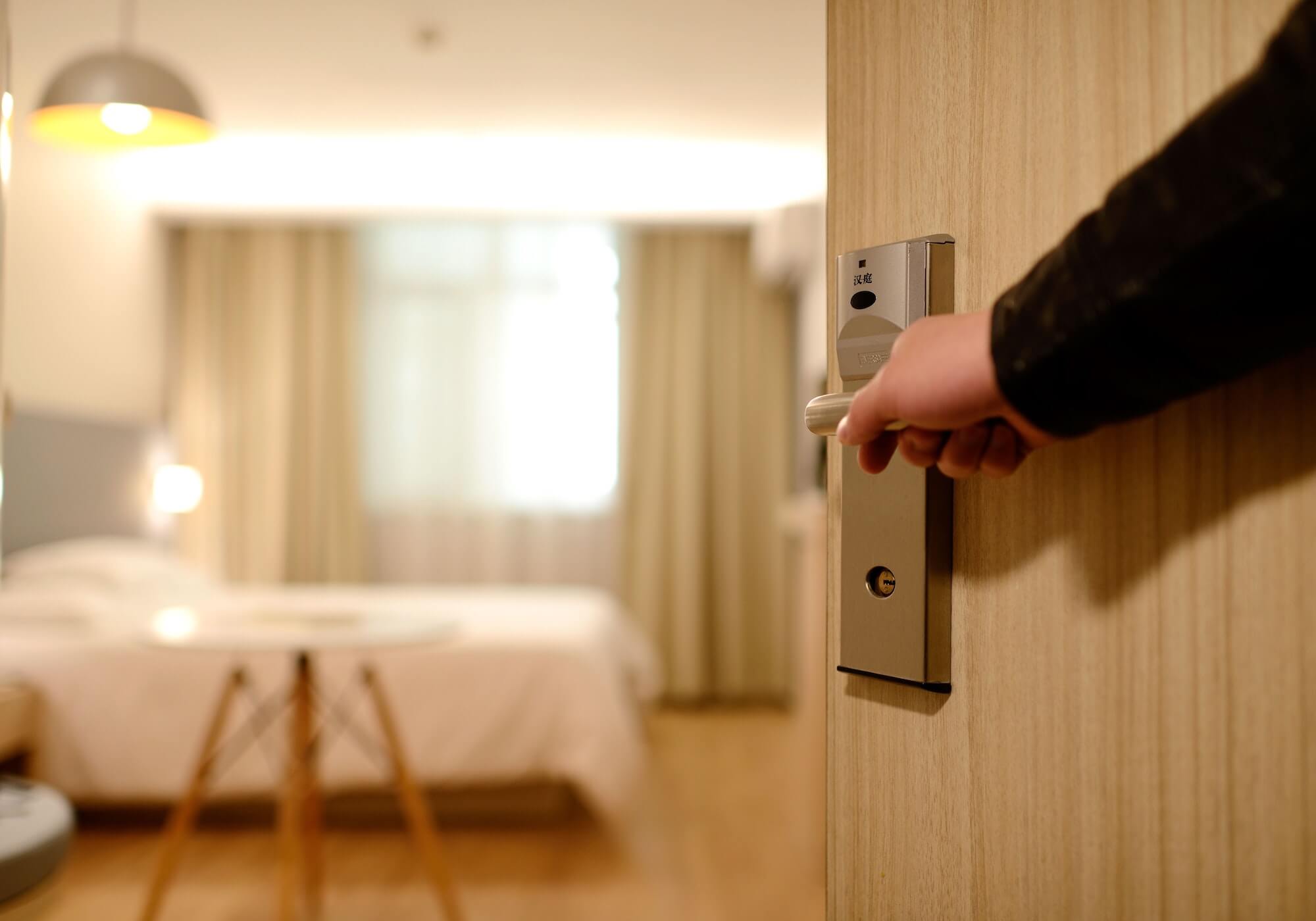 Hand opening wooden hotel room door, with out of focus room visible behind.