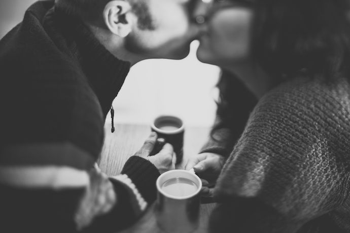 black and white close up photo of man and woman kissing while holding mugs of coffee