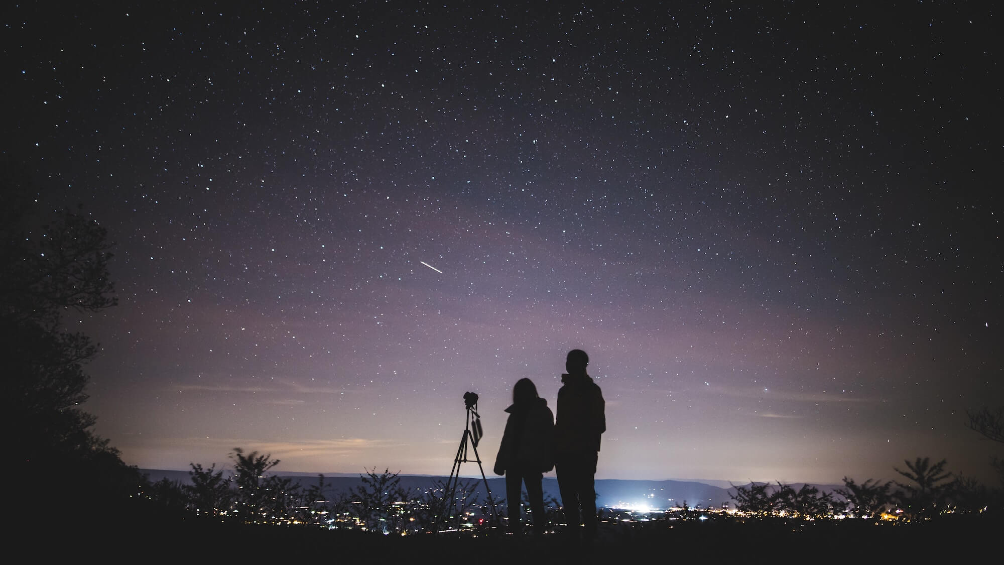 Silhouette of man and woman standing on a hill against a starlit sky.
