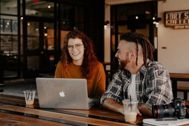 A woman with red hair and glasses and a man with dreadlocks, laughing in a cafe with coffee and a MacBook