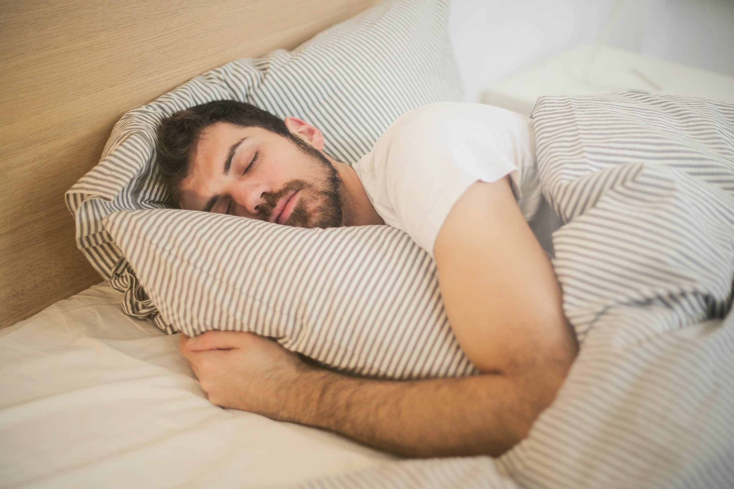 Bearded man sleeping in bed with stripy white sheets.