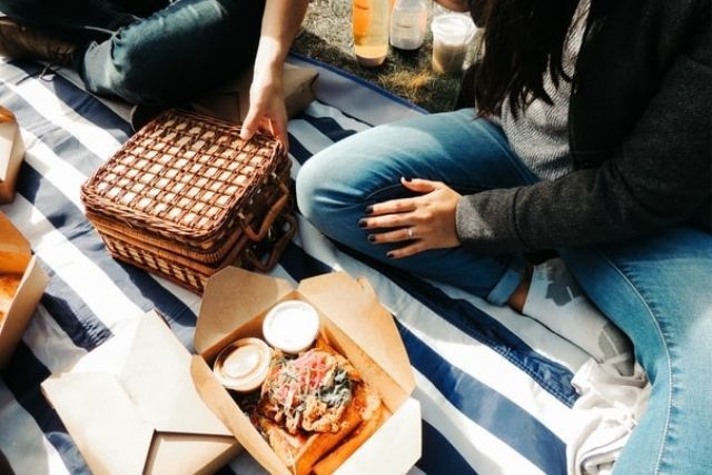 Takeaway food and picnic hamper on blue and white striped picnic blanket, with a man and woman sitting beside 