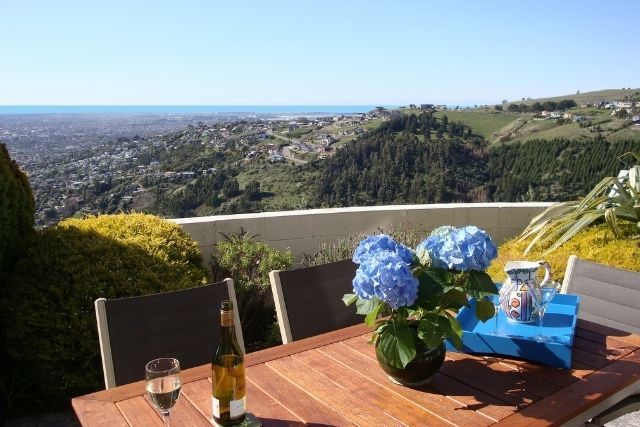 Outdoor wooden table with a bottle of wine and a vase of blue hydrangeas, looking out over the view of Christchurch and the Pacific Ocean 