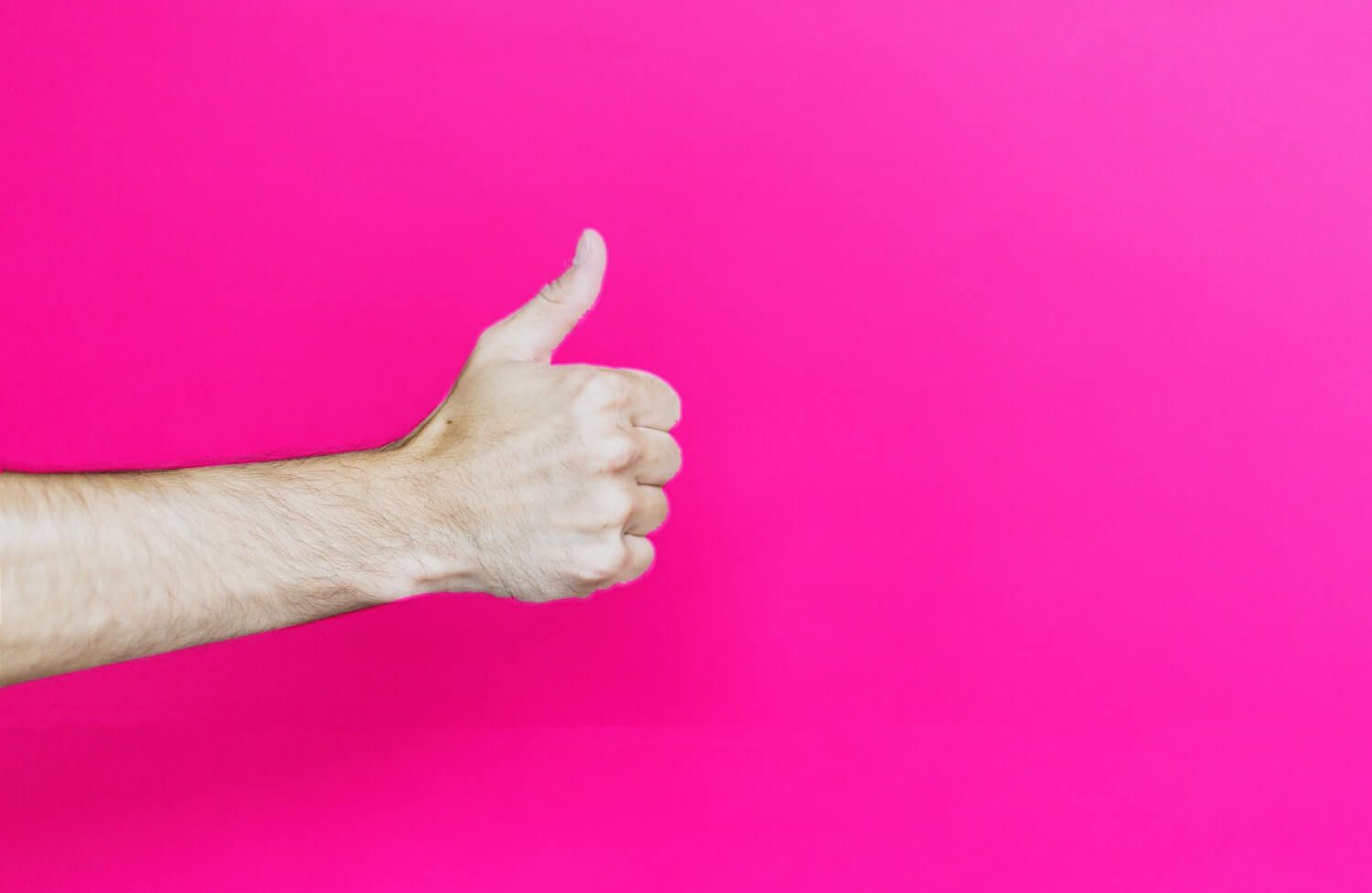 Male arm making thumbs up gesture against a bright pink background