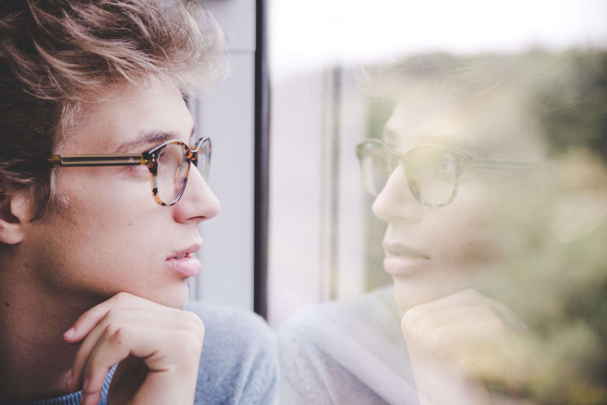 Man with glasses looking out of window with his reflection visible in the glass.