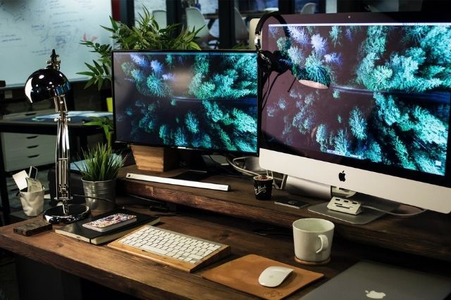 Tidy desk set up, with chrome lamp, diary, mug, and two screens with a forest view screensaver