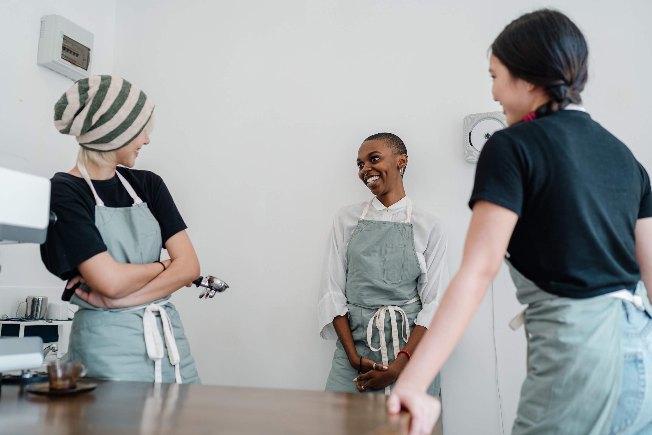 Three female baristas wearing matching green aprons and laughing. 