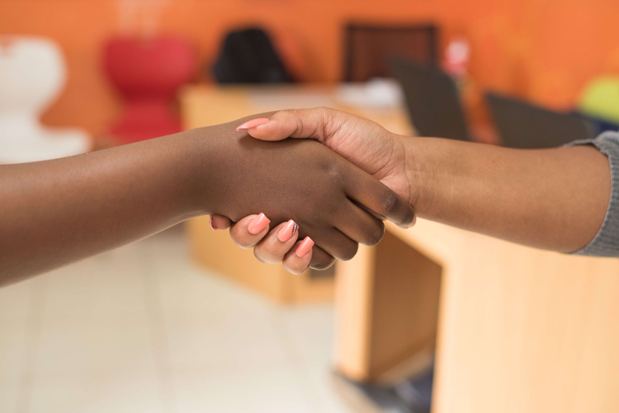 Close-up photo of two people shaking hands: hand on the left is dark-skinned; hand on the right it lighter skinned and has long fingernails painted pink.