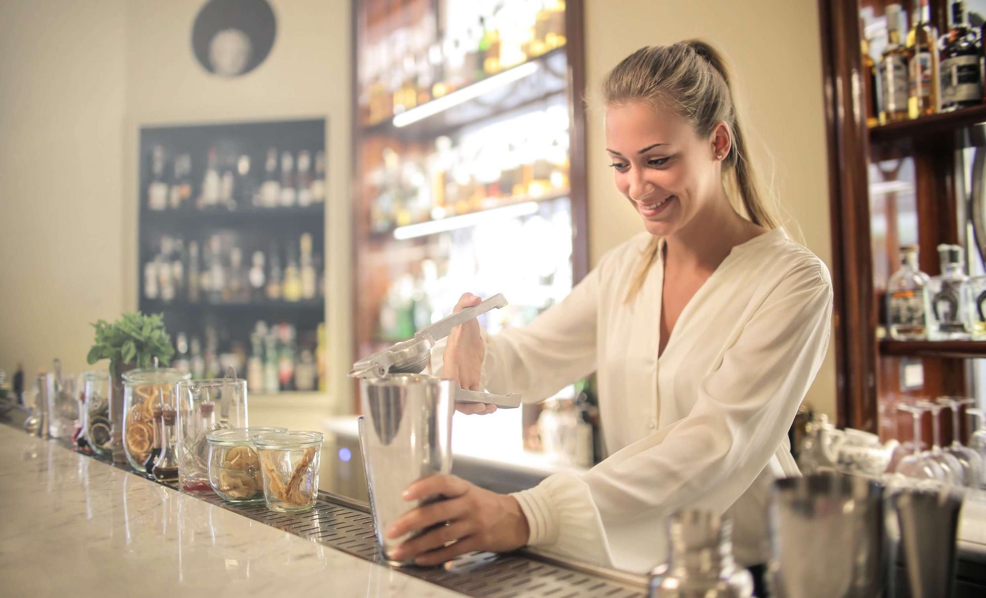 Smiley, blonde-haired female bar tender making a cocktail behind a bar well-stocked bar.