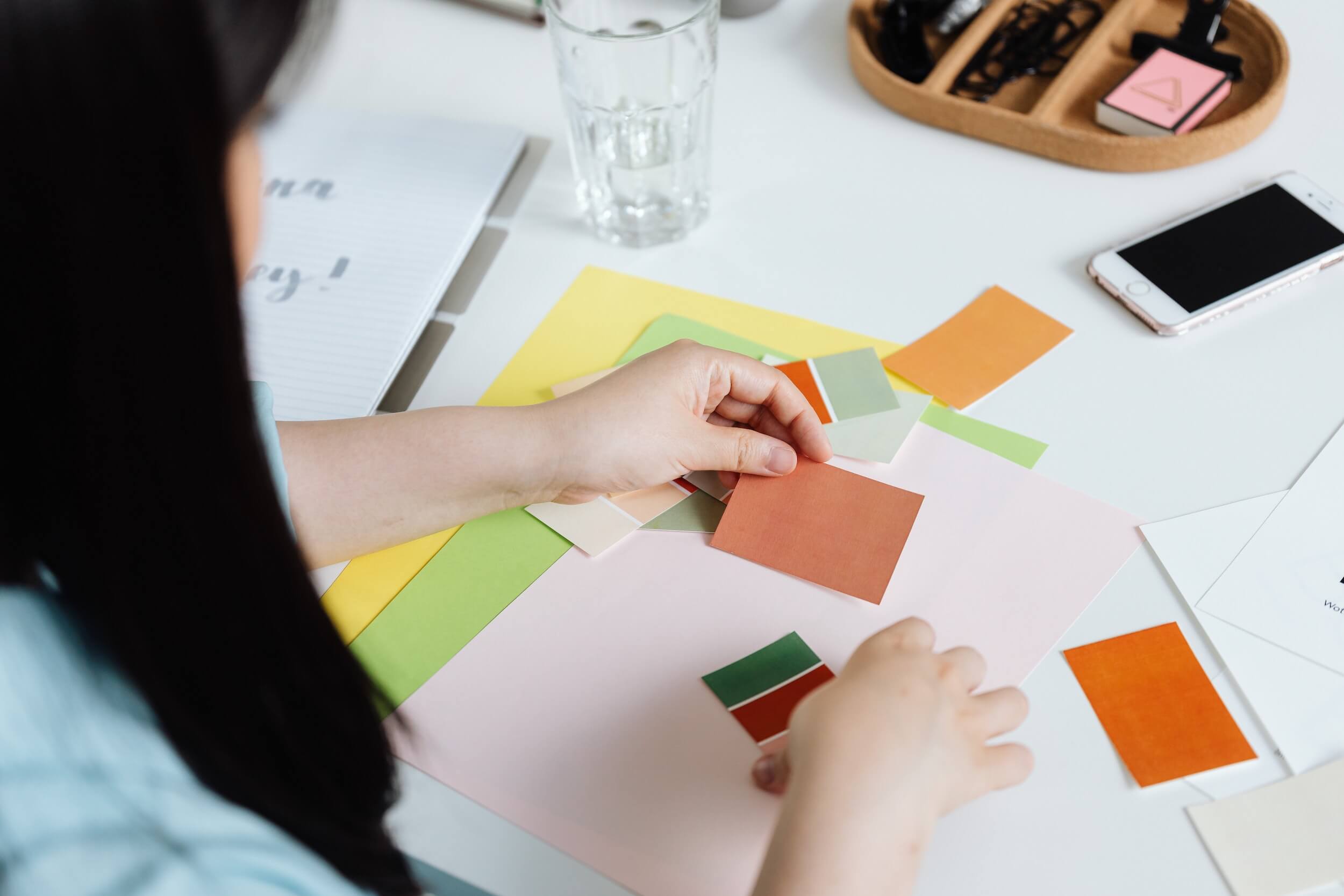 Woman with black hair studying different coloured card samples on a white desk.