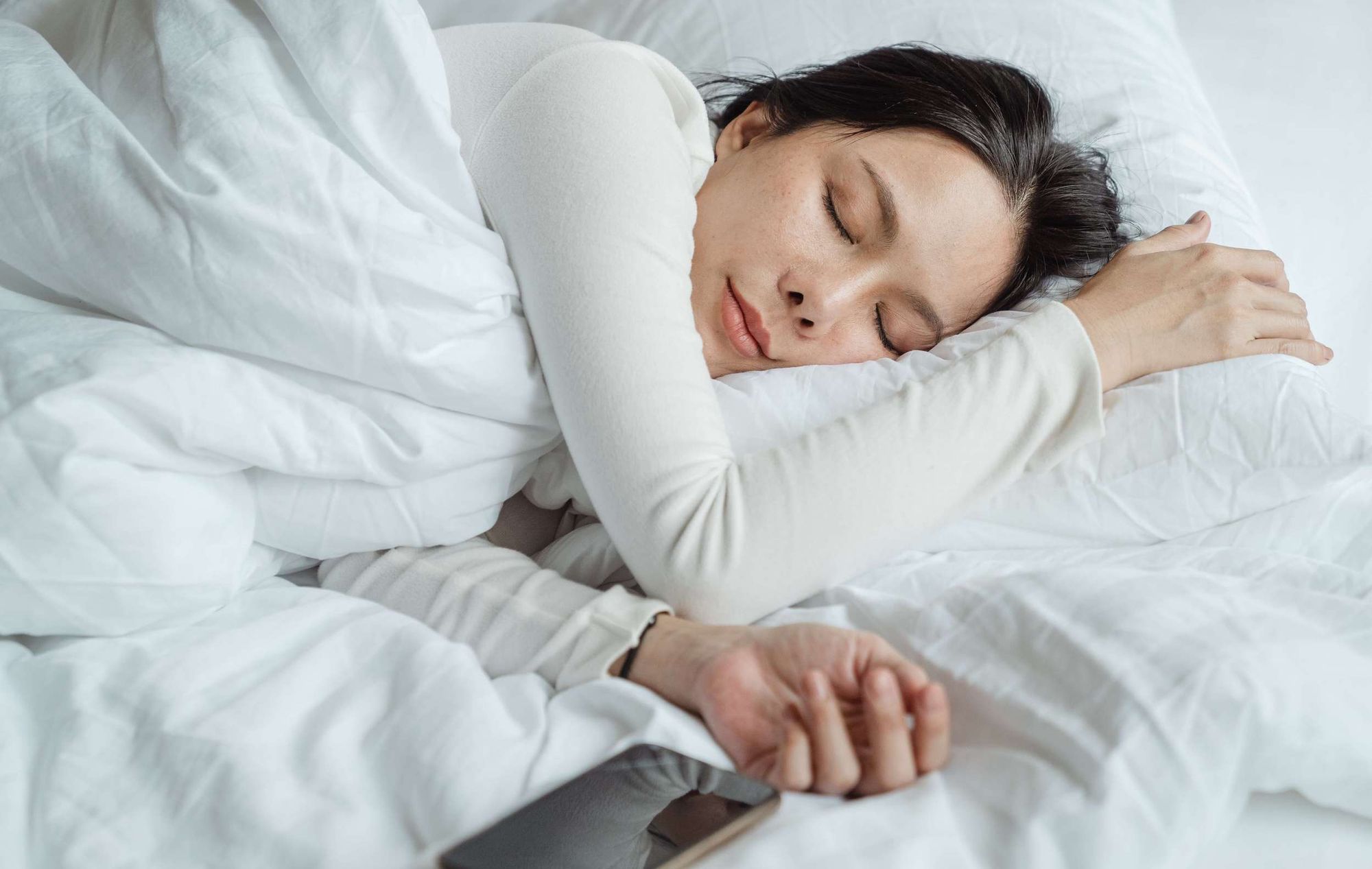 A woman with dark hair sleeping on white bedsheets next to her mobile phone.