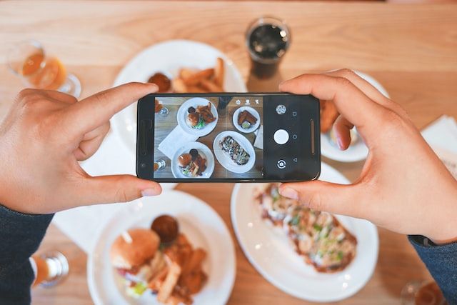 Man's hands holding phone, taking a landscape photo of plates of food set on a wooden table