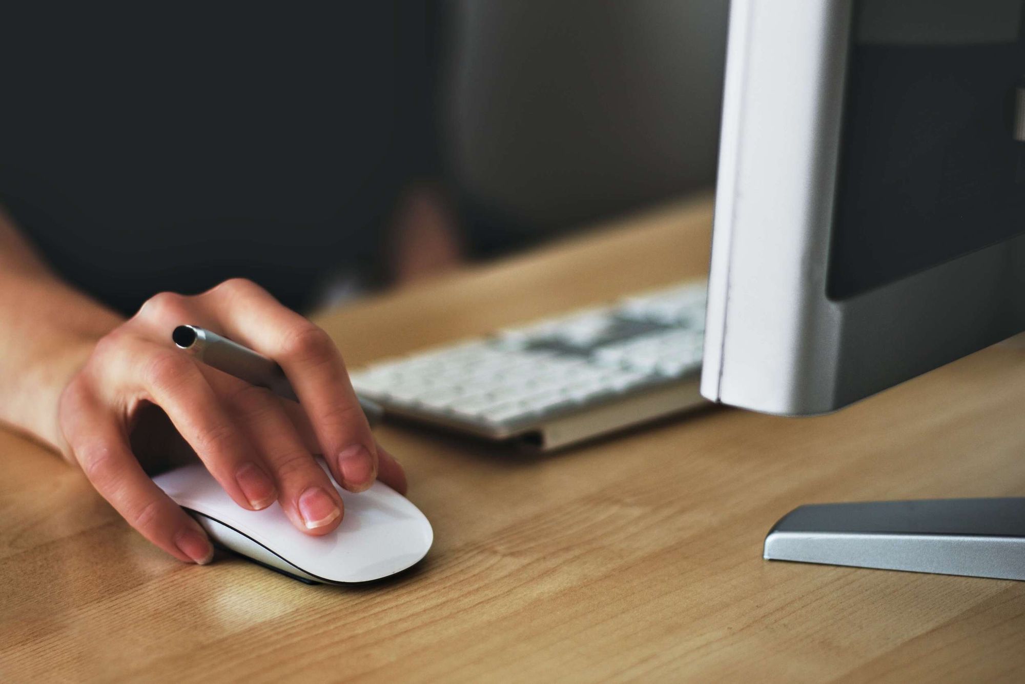 A hand with a pen between two fingers operating an Apple mouse on a wooden desk.