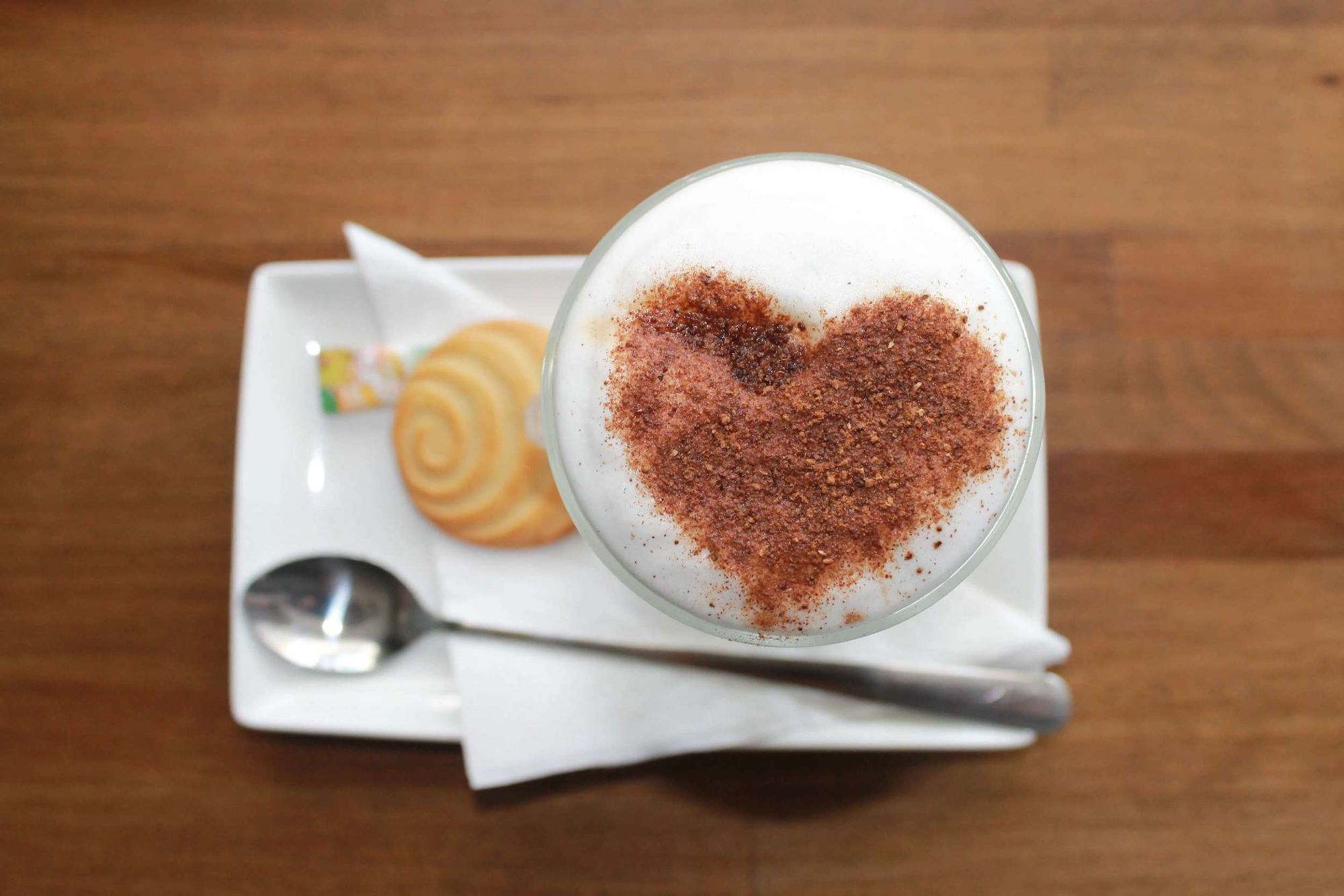 A latte coffee with a heart shape in cinnamon on the froth, alongside a cookie and spoon. 