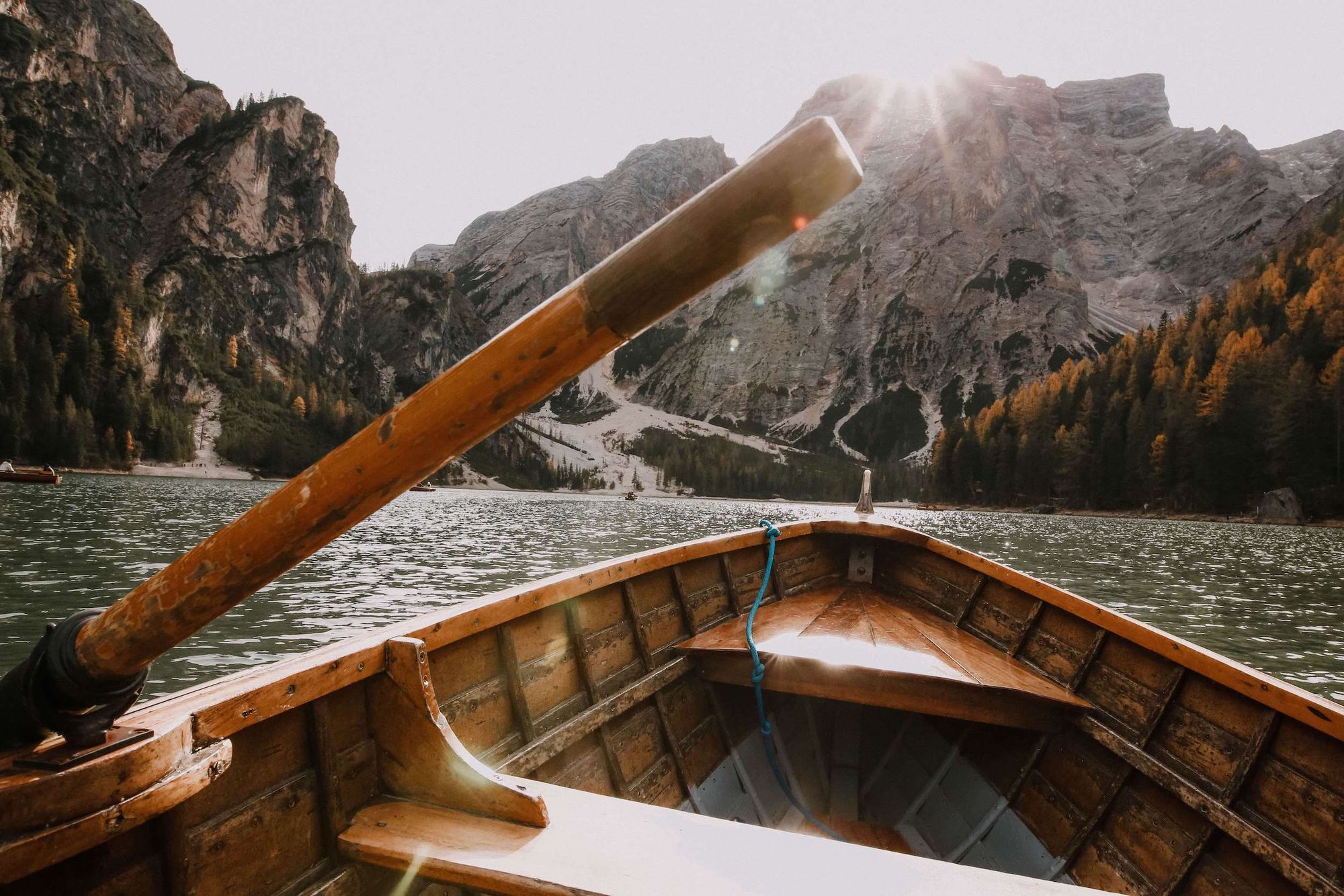 Wooden row boat with one oar visible on a calm lake surrounded by snowy mountains and woodland.