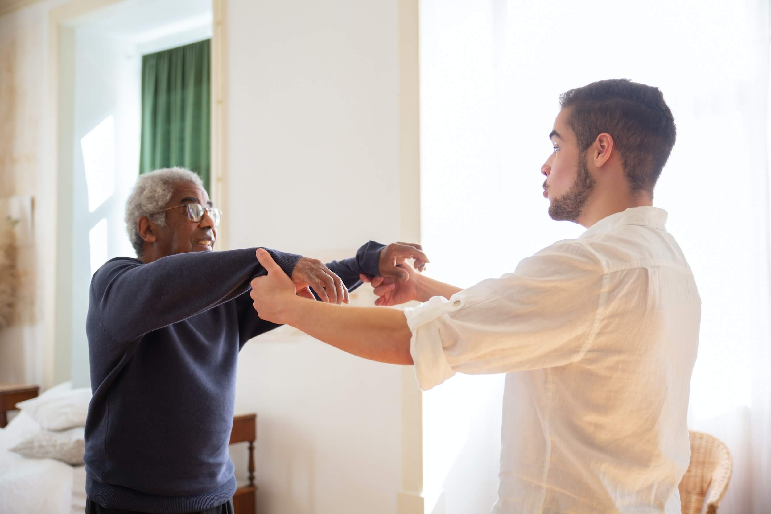 Care assistant wearing a white shift raising the arms of an older service user.