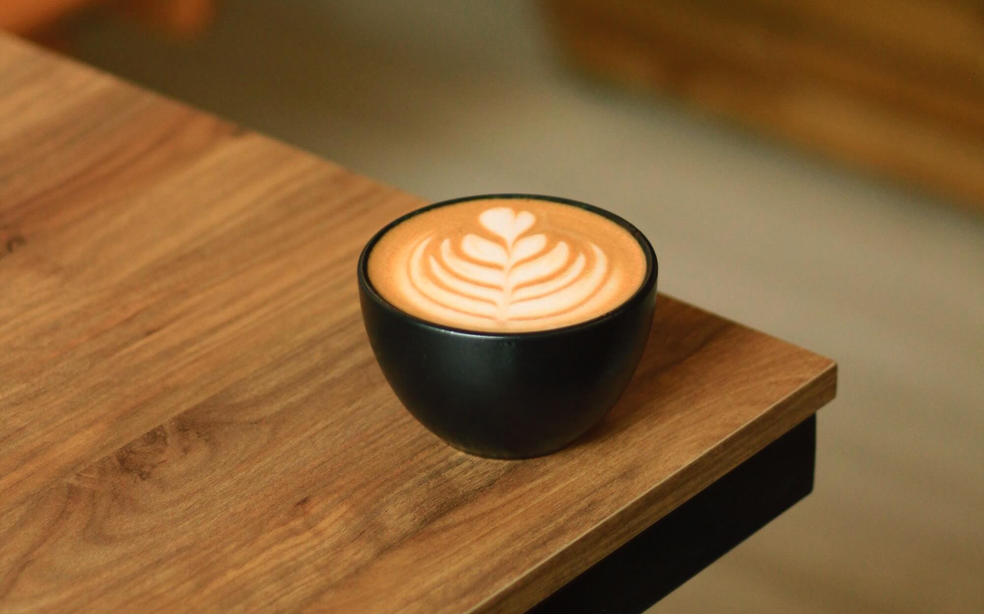 A pumpkin-spiced latte in a matte black cup on the corner of a wooden table.