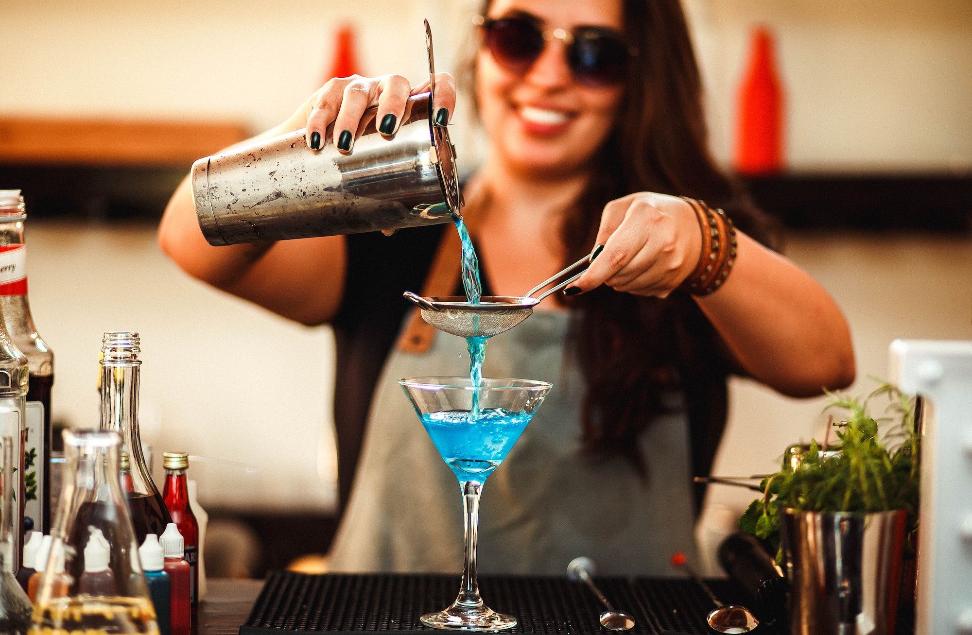 Bartender with long hair and sunglasses pouring a blue liquid from a cocktail shaker into a glass.