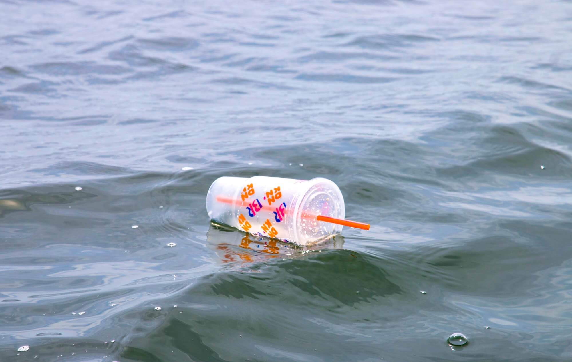 An empty plastic takeaway cup with red straw floating in water outdoors.