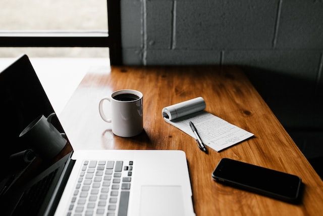 Macbook, phone, notepad and pen, and a white mug of black coffee on a wooden table