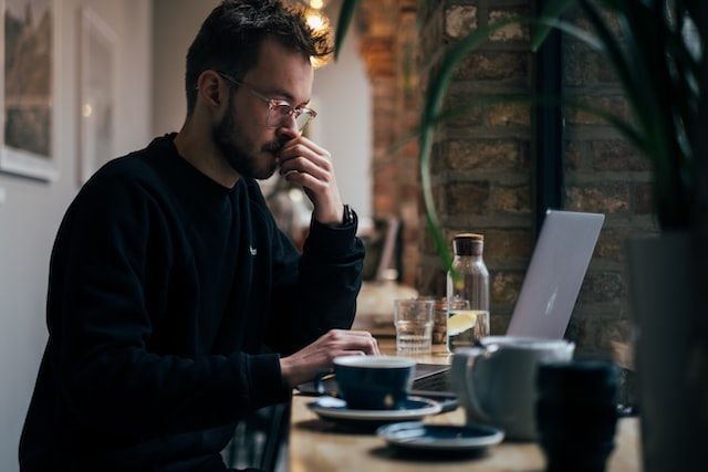 Man in a cafe looking worried and staring at his laptop screen, coffee cups in foreground
