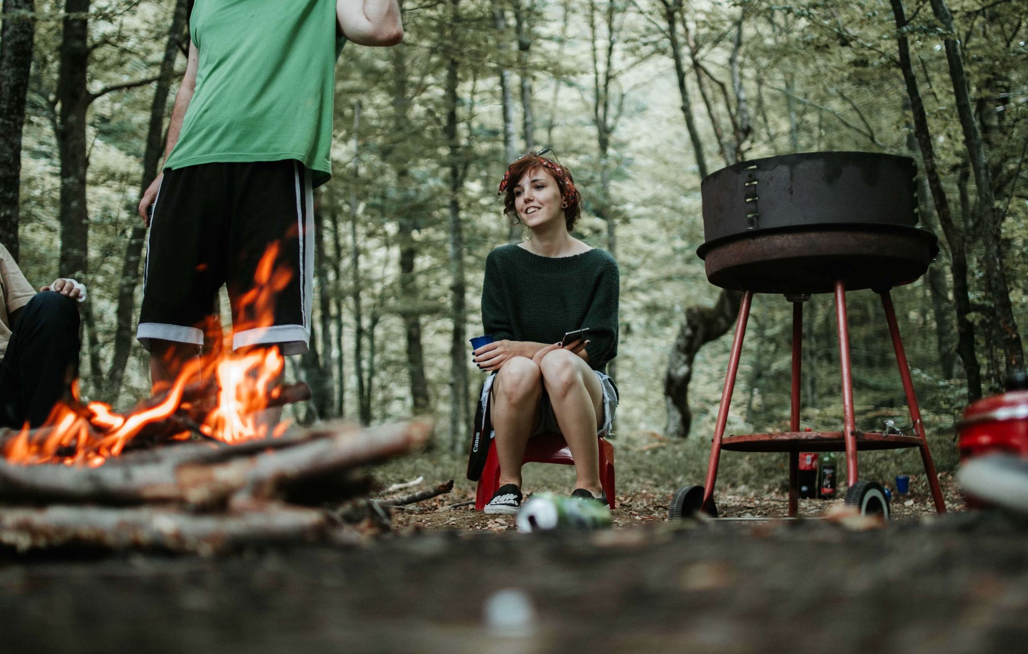 Young woman sitting on a camping stool in a forest in front of a campfire