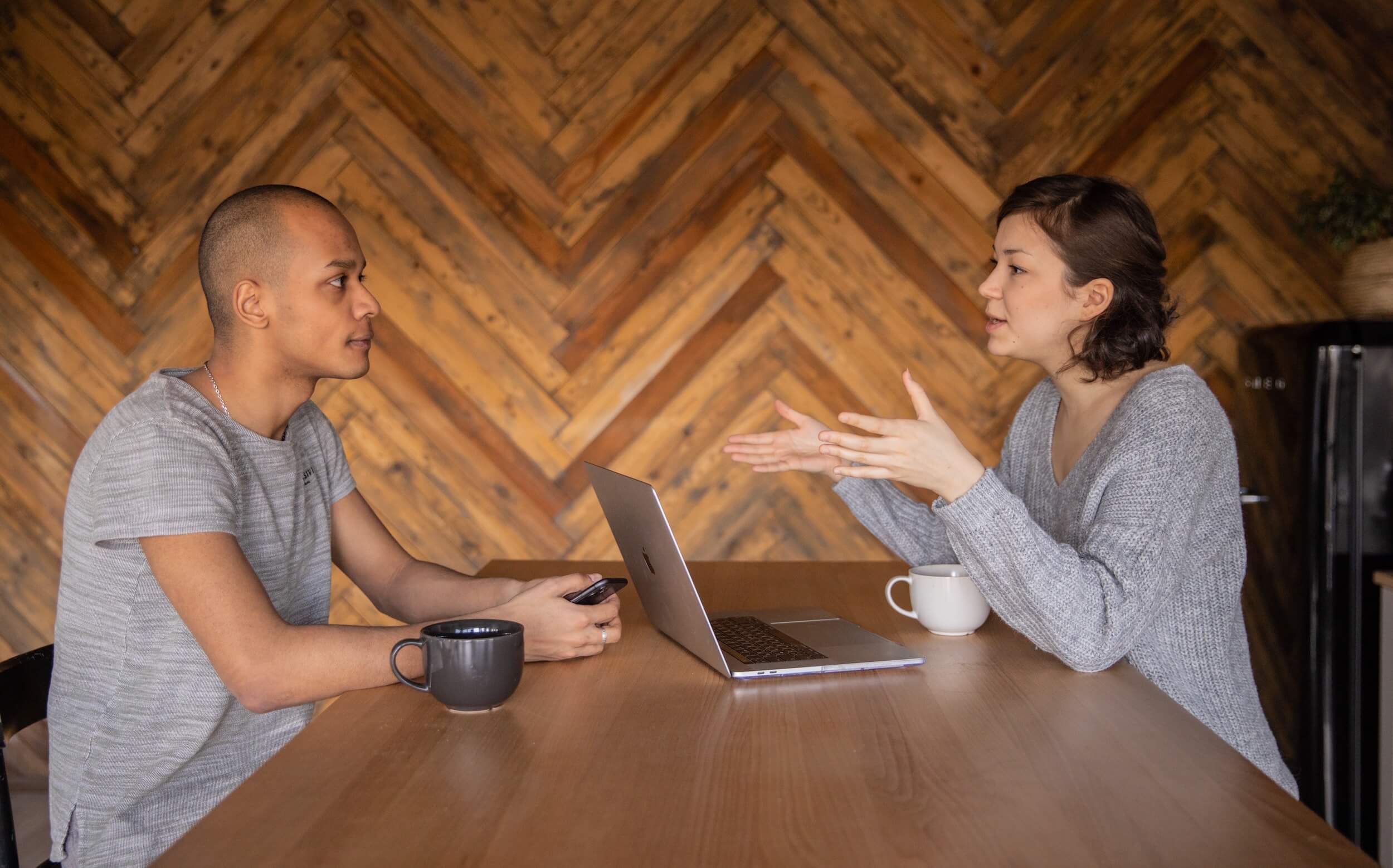 Man and woman talking across a table with a wood panel wall behind them.
