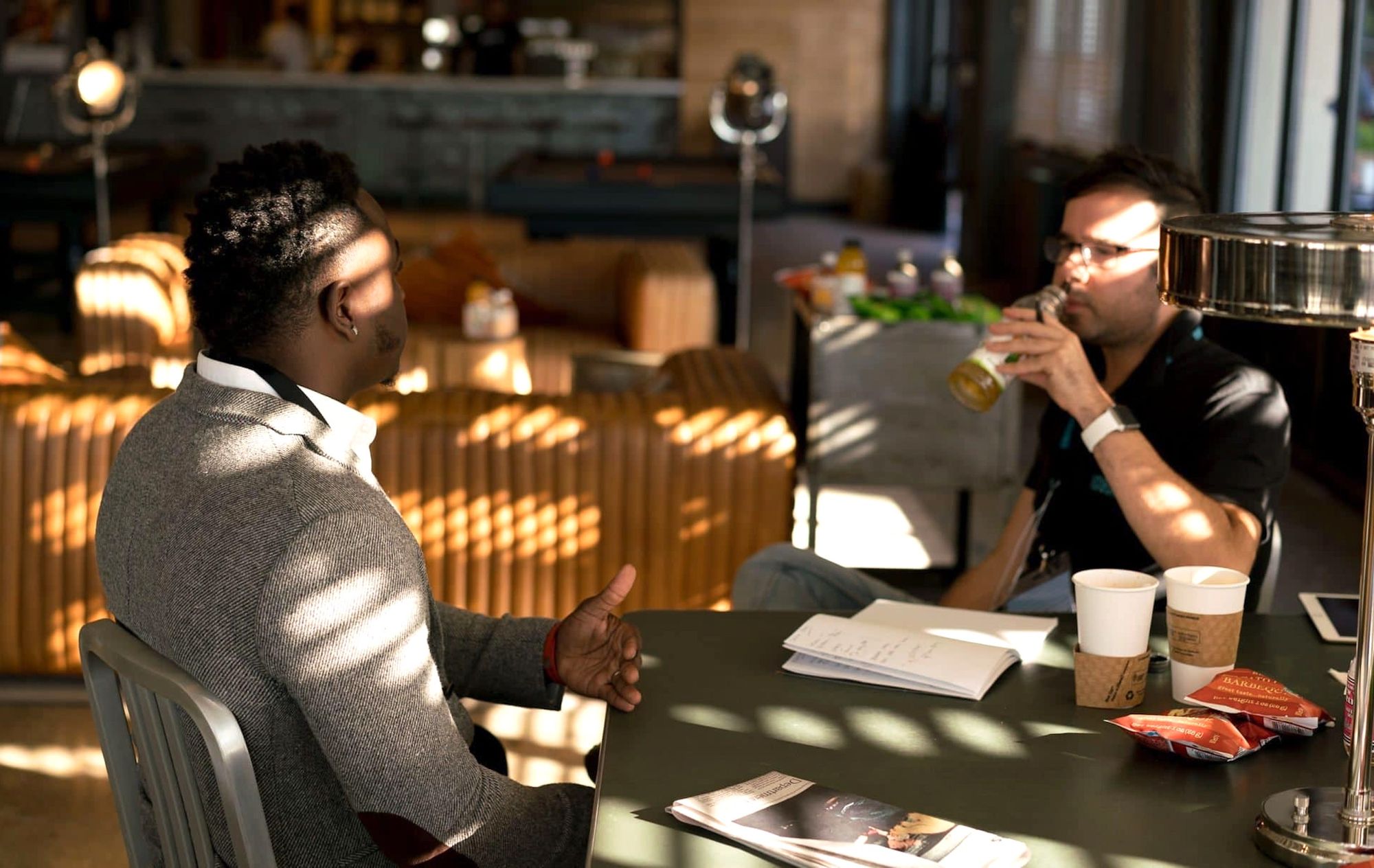 Two men talking while seated at a table covered in empty coffee cups.