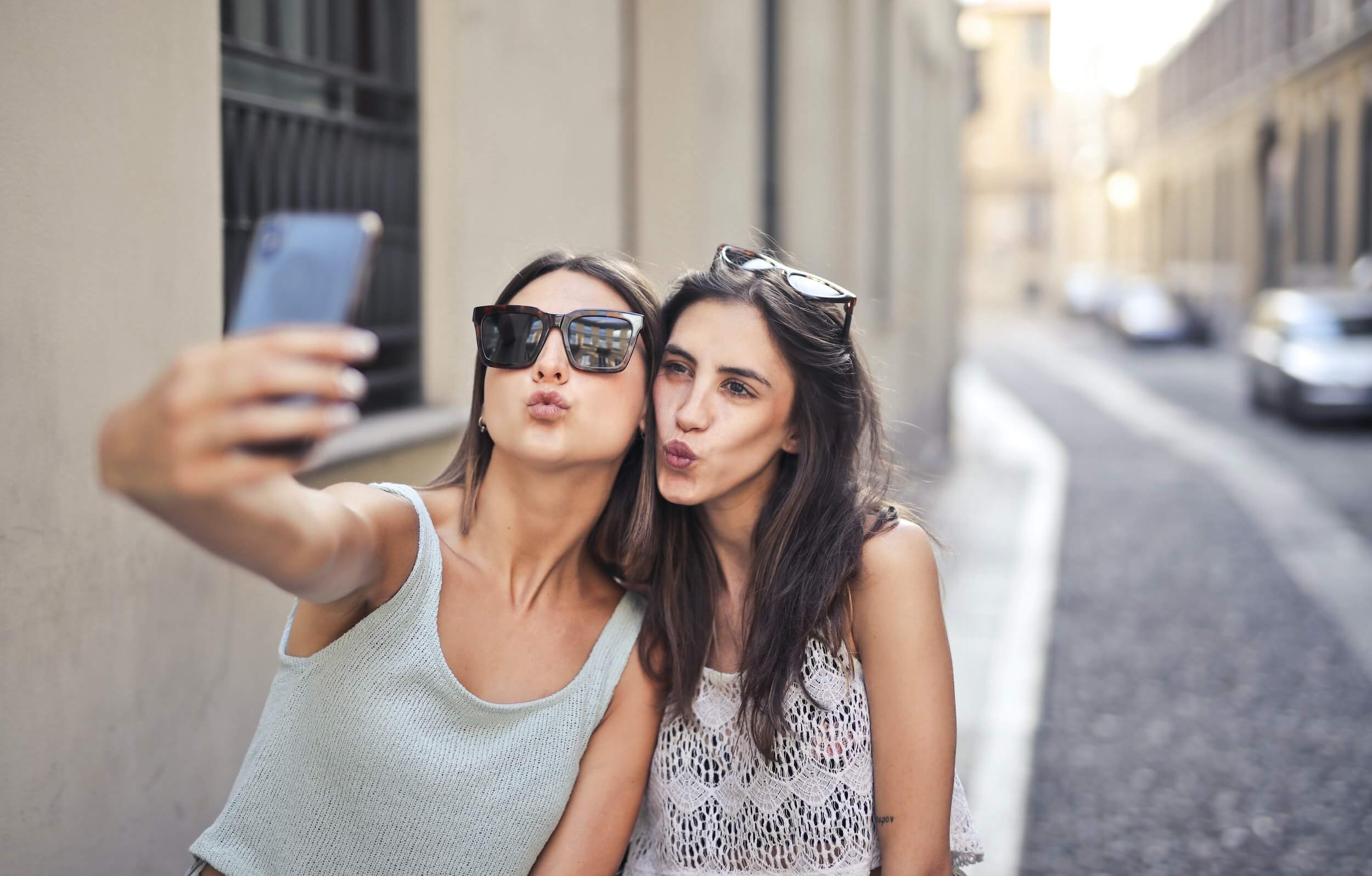 Two women wearing sunglasses taking a selfie on a street with cars in the background