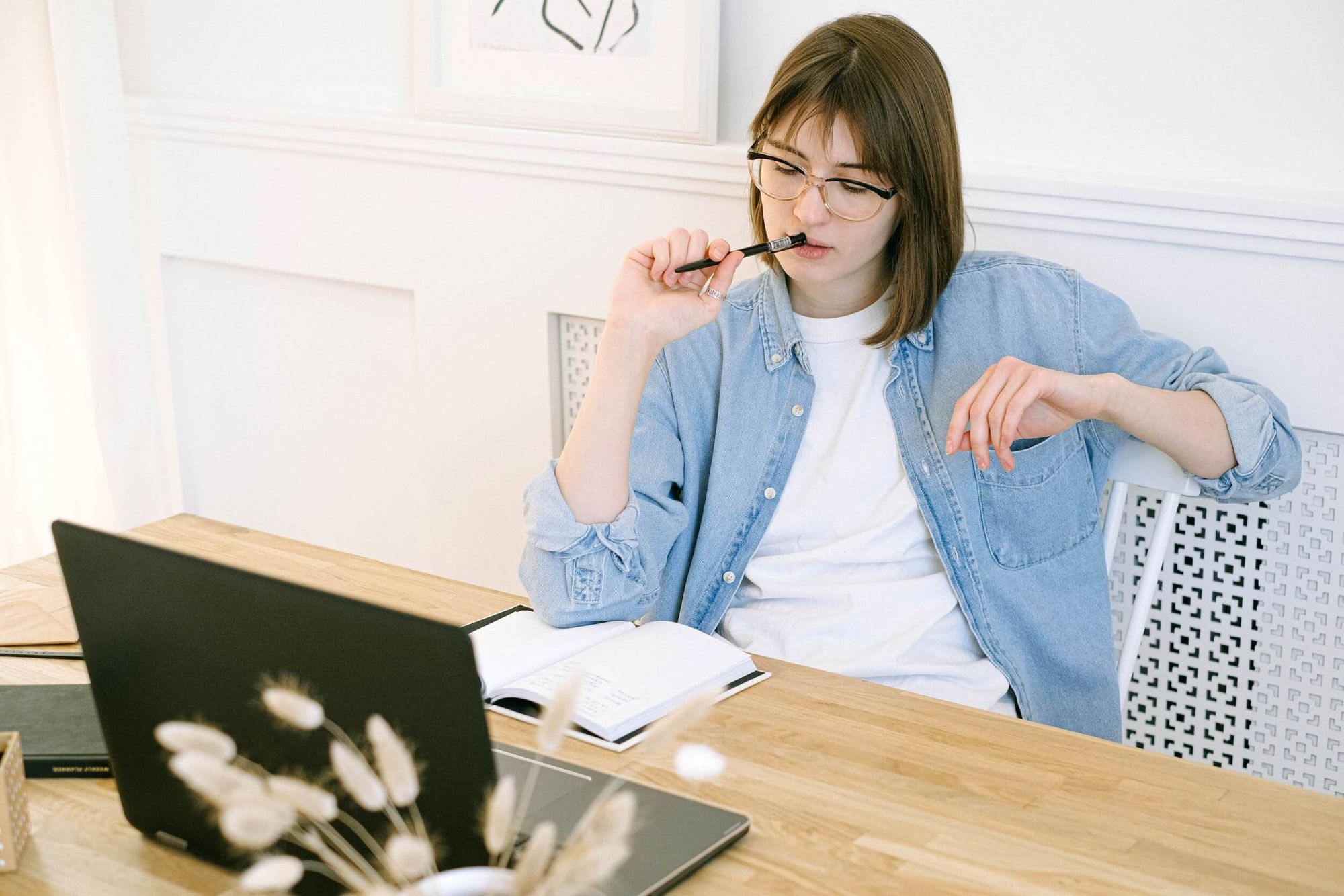 Woman wearing a blue shift thinking while sitting at her desk with notebook and laptop