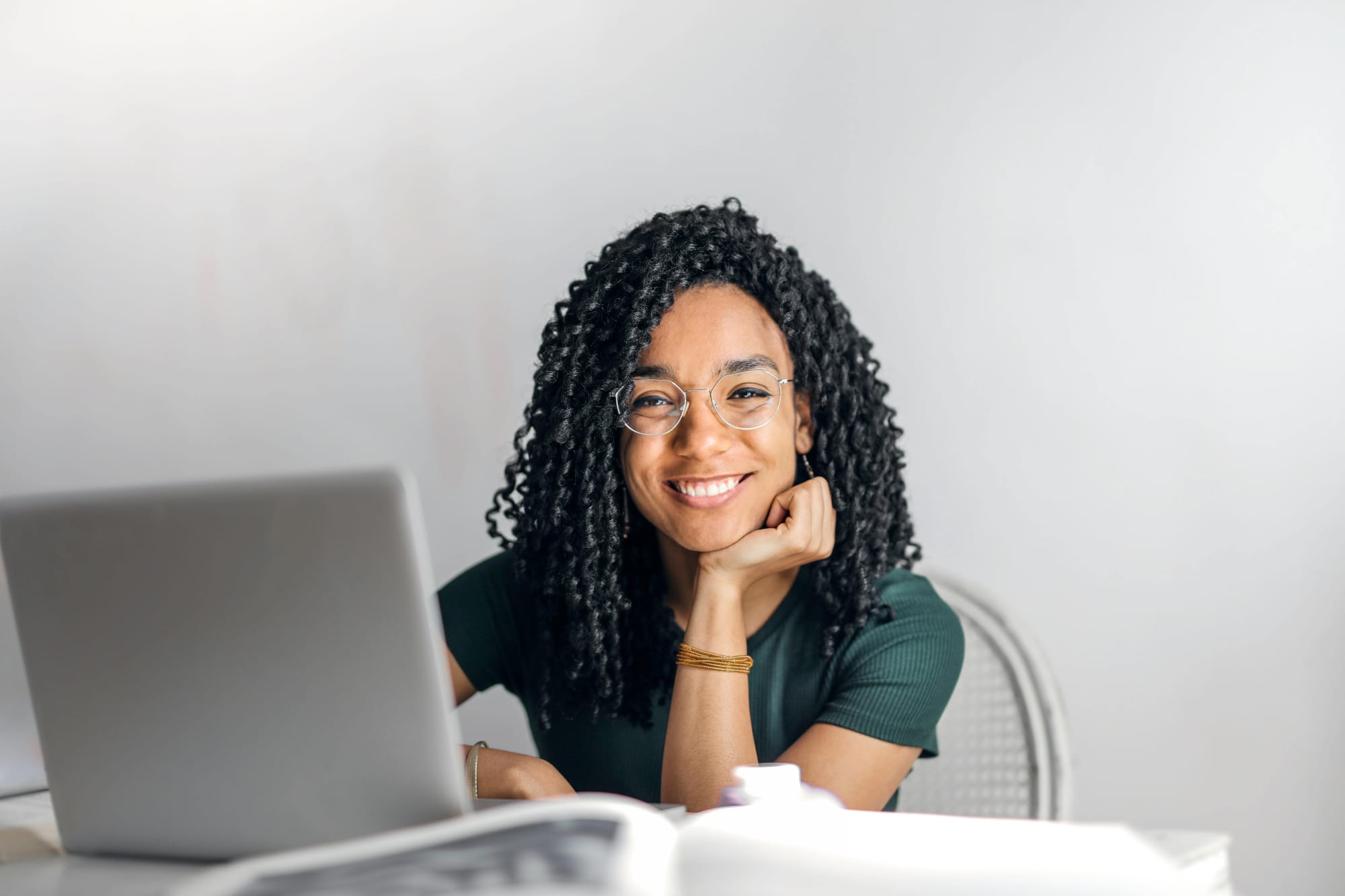 A woman sitting a a table with a laptop, leaning her chin on her hand and smiling at the camera.