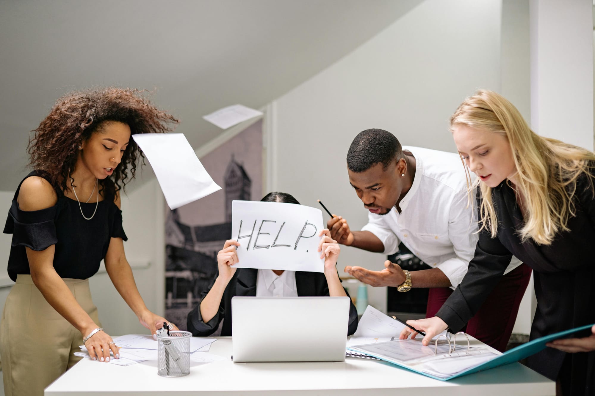Four frustrated office workers surrounding a table, paper flying around them. One person hides their face behind a paper that reads: 'HELP.'