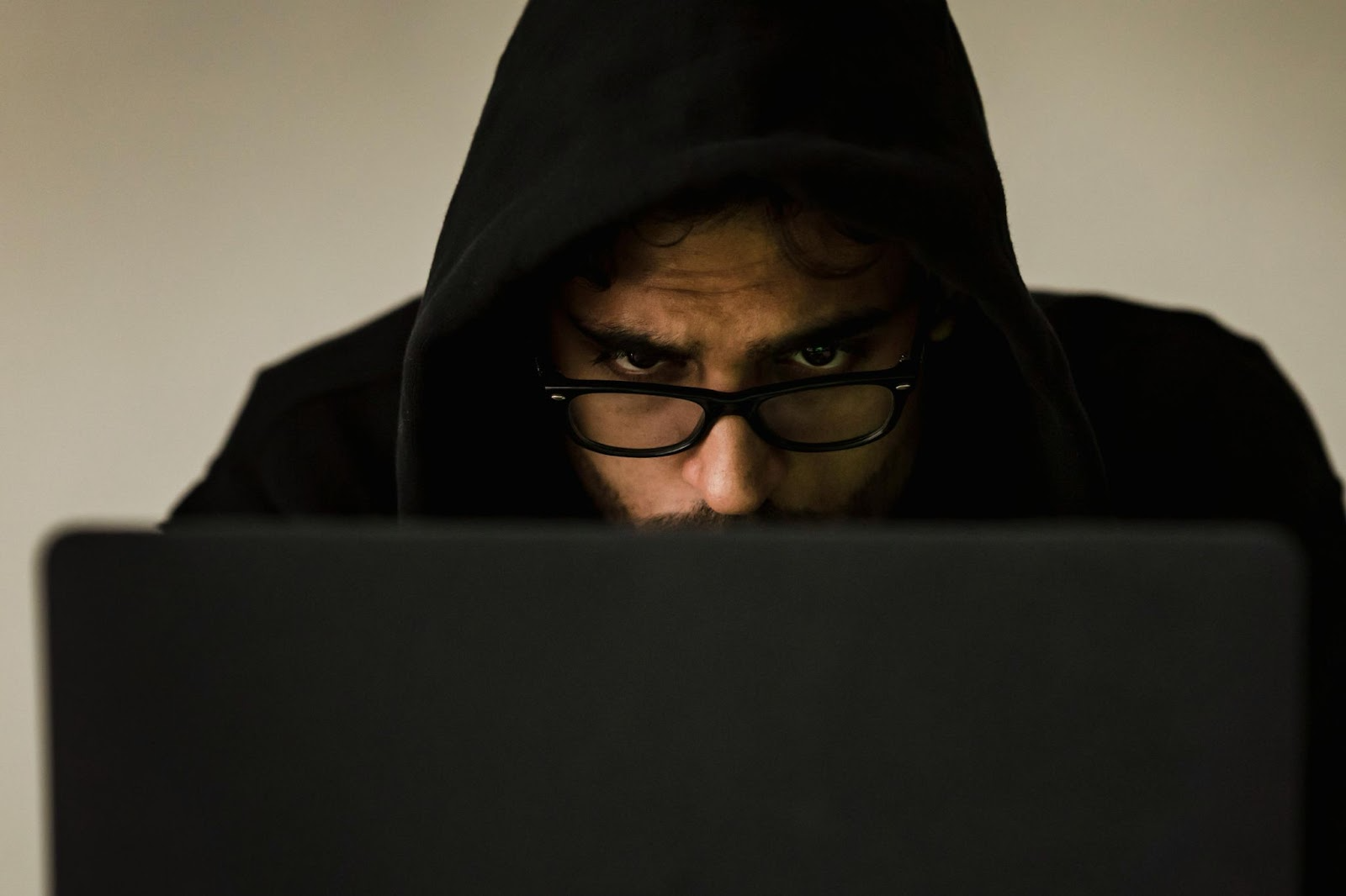 A hooded man in glasses looking fiercely at his laptop screen.