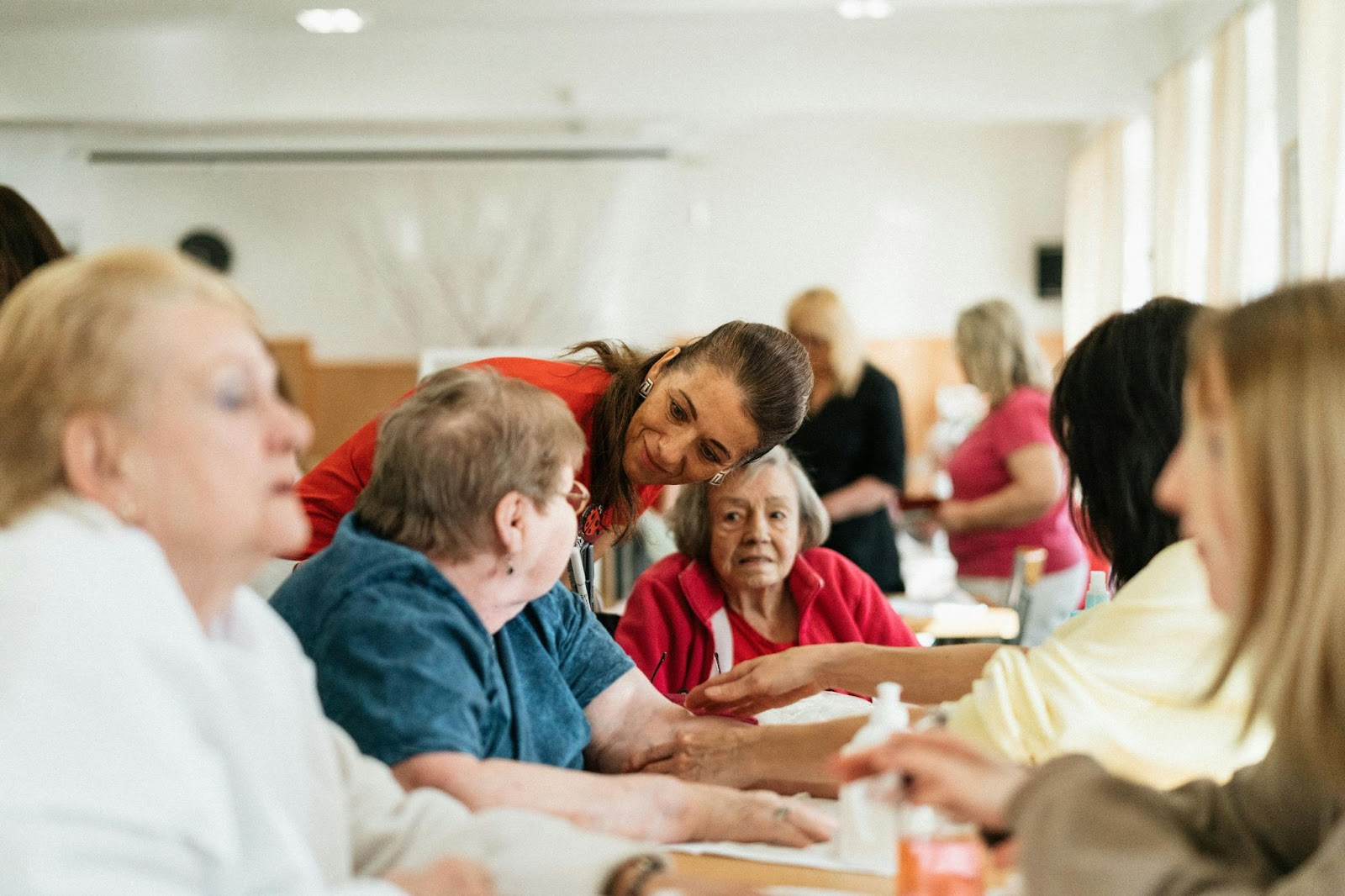 A group of elderly patients gathered around a table. A carer is bent between them, listening to one.