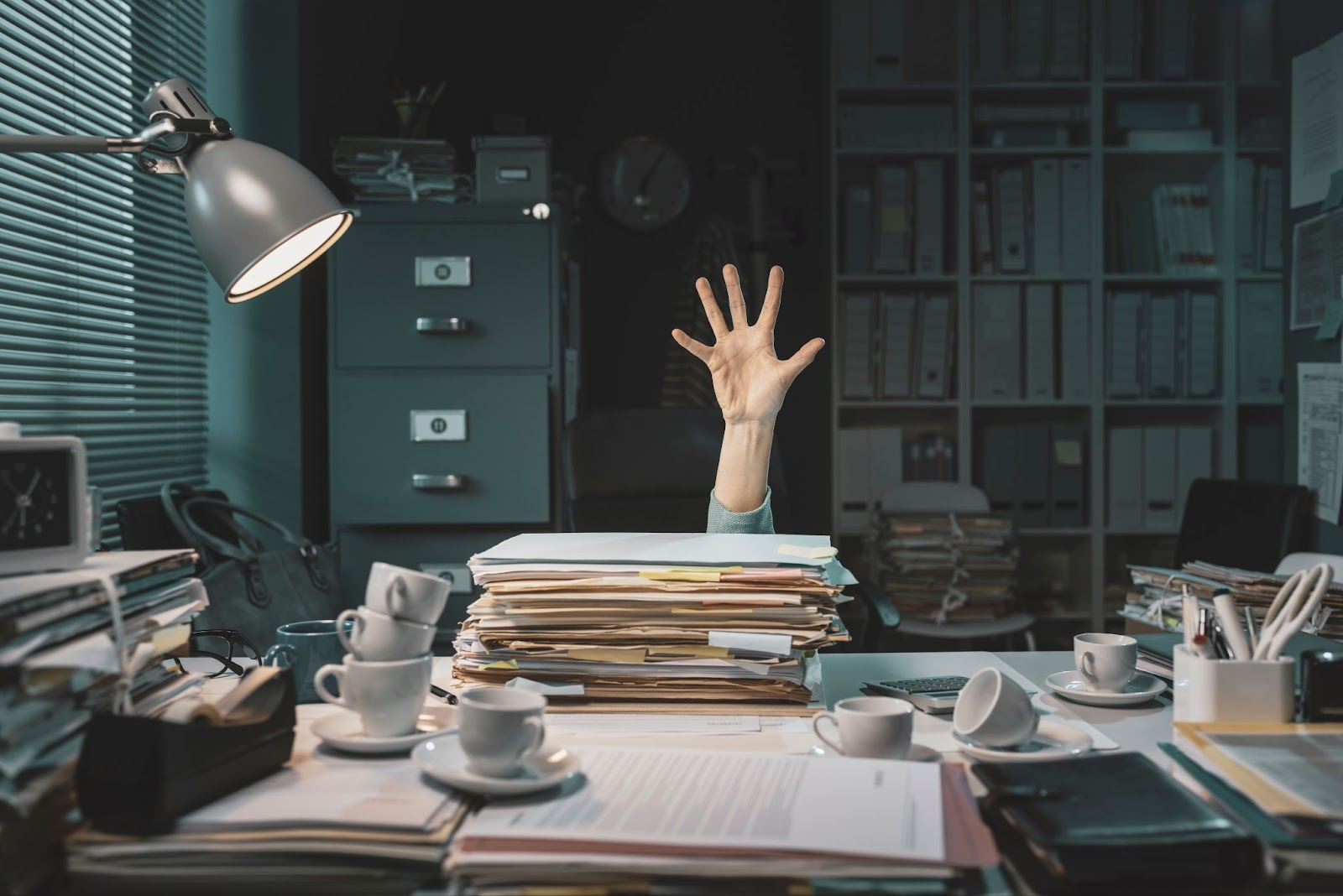 A grey office, files and cabinents in the background, with a desk covered in stacks of paperwork and empty coffee cups. A hand reaches out from behind it all, as if for help.