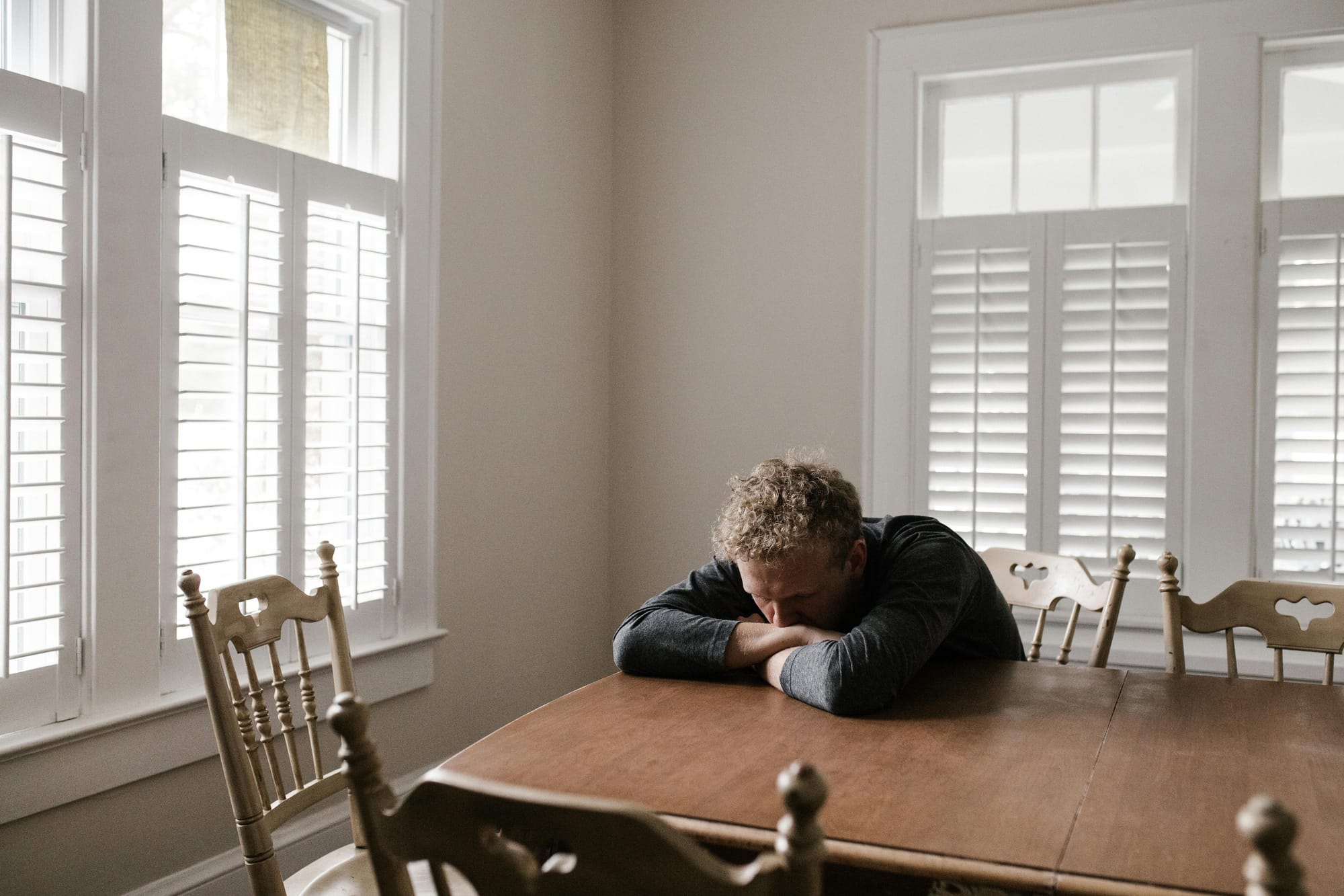A man leanining his head into his crossed arms on a dining room table.