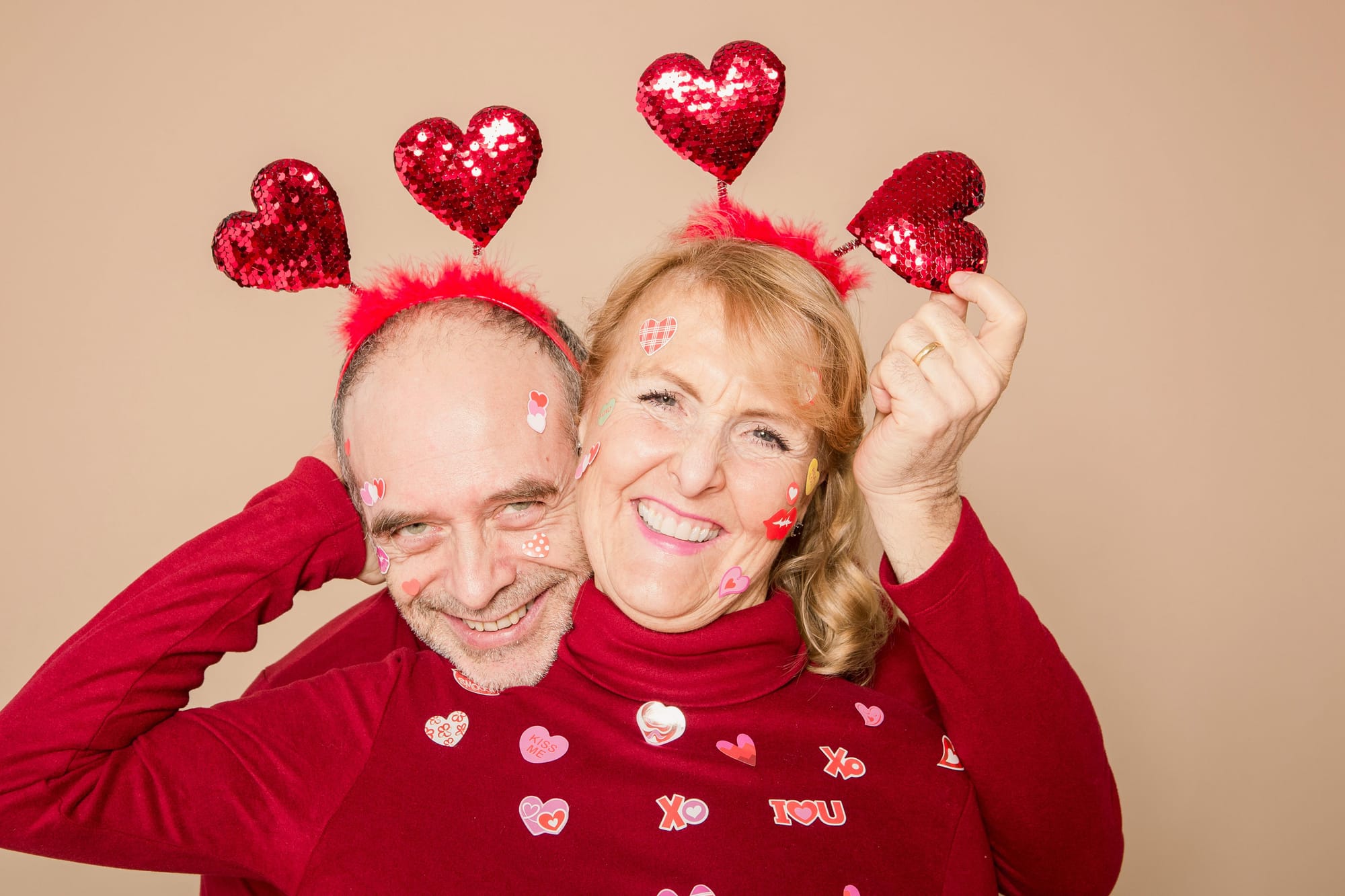 A couple are dressed in red, wearing sparkly red heart headbands. Heart stickers cover their faces and clothes as they pose cheek-to-cheek for a fubthe photo.