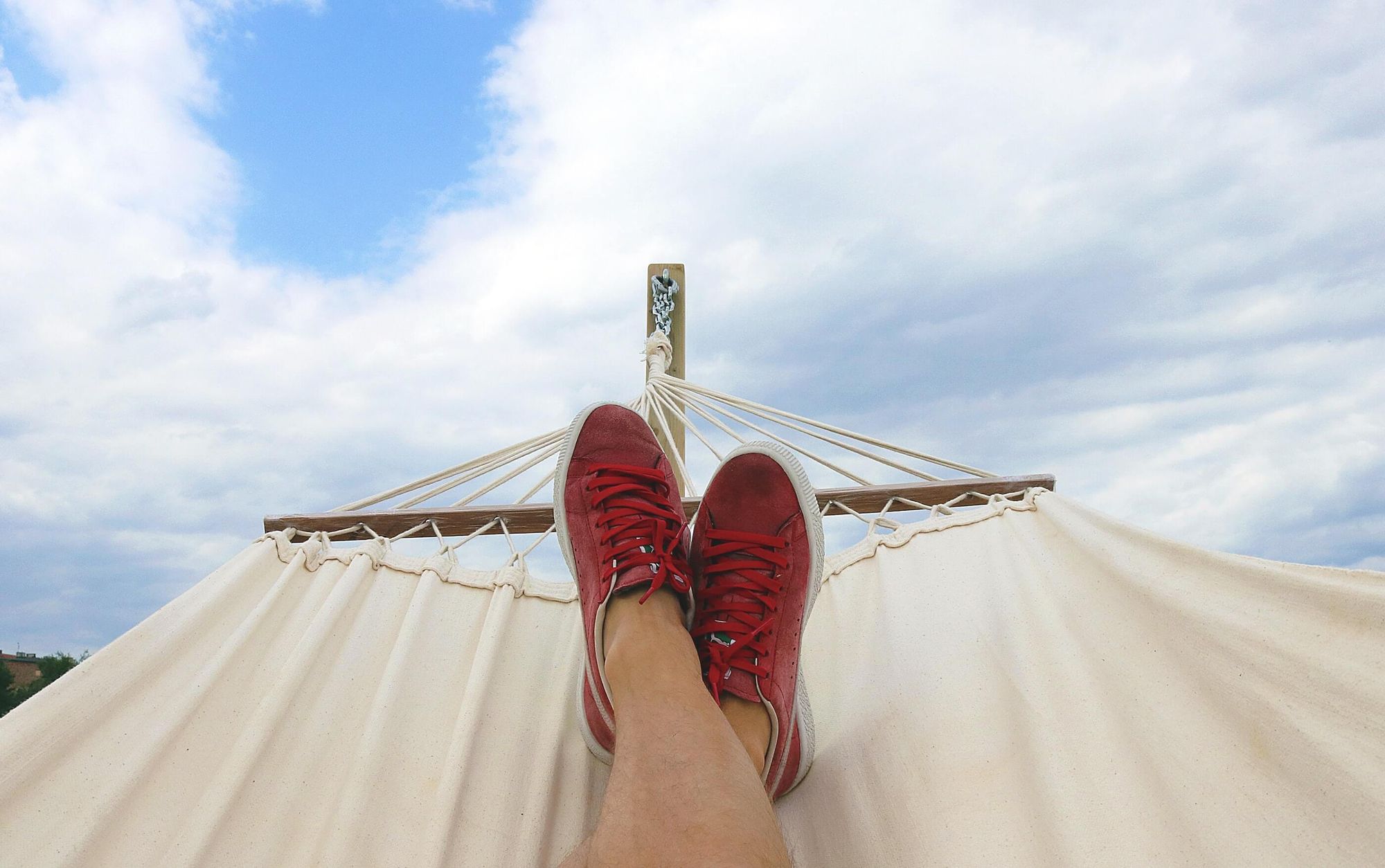 A pair of feet wearing red deck shoes in a canvas hammock with blue sky in the background.