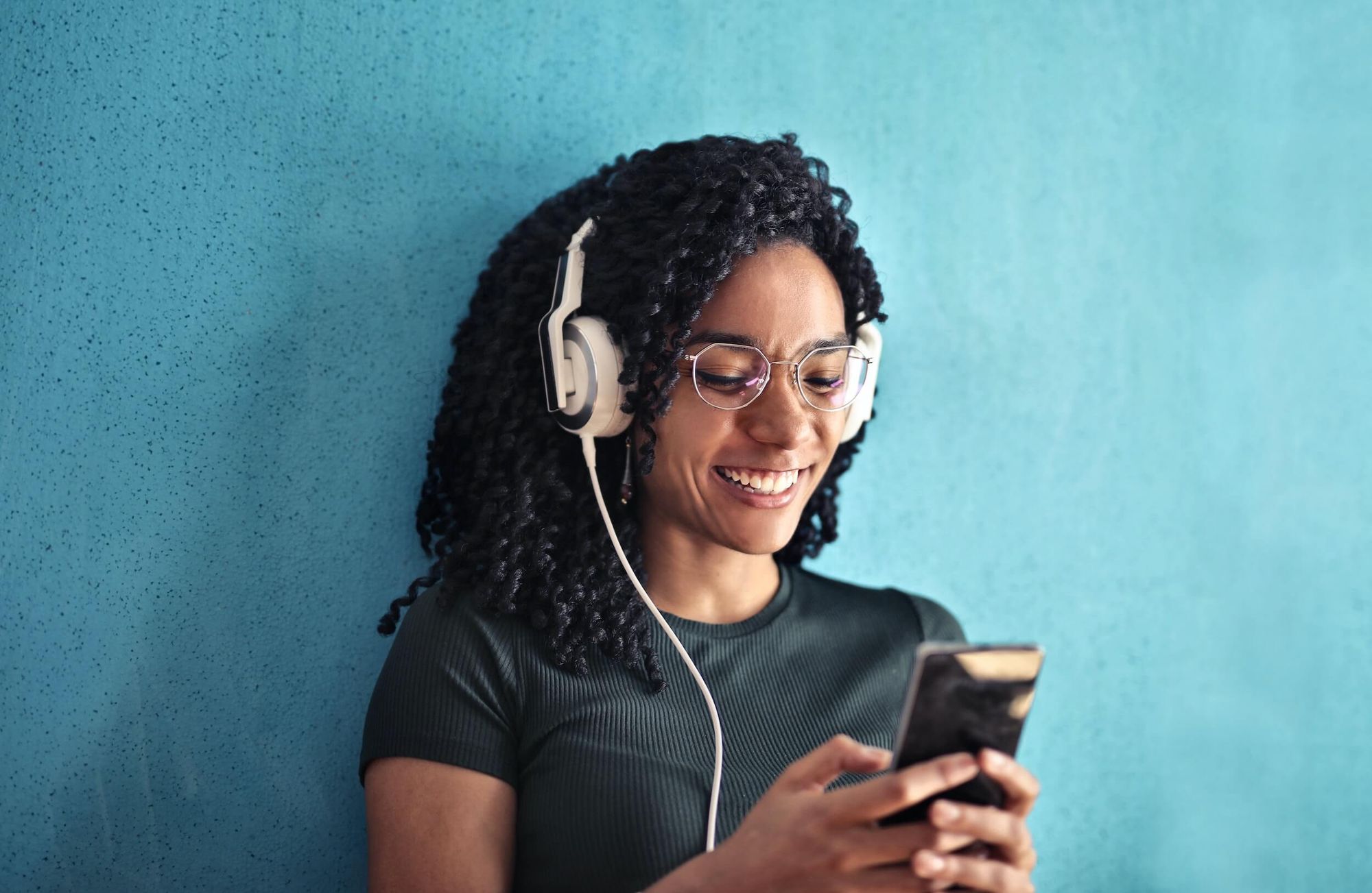 Woman of colour smiling while wearing glasses and large white headphones, looking at a mobile phone.