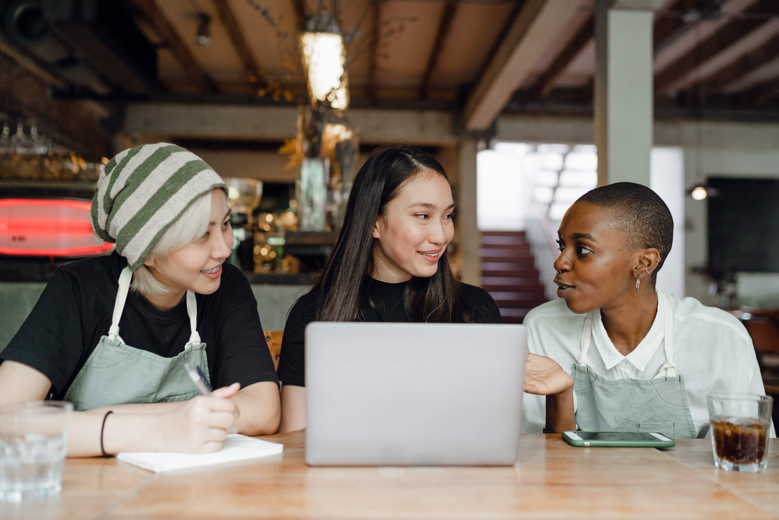 Three female baristas in conversation while sitting at a desk with a laptop computer.