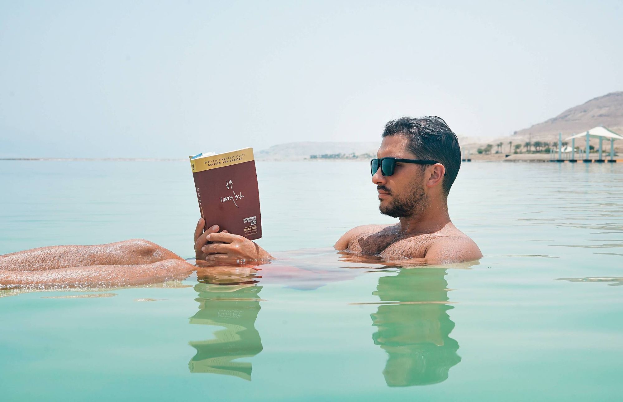 Man wearing sunglasses floating in the sea reading a book.