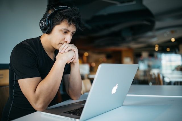 Man wearing headphones, staring at an Apple laptop, and looking slightly anxious