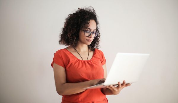 Woman with dark, curly hair and glasses holding a silver laptop computer standing in front of a white background.